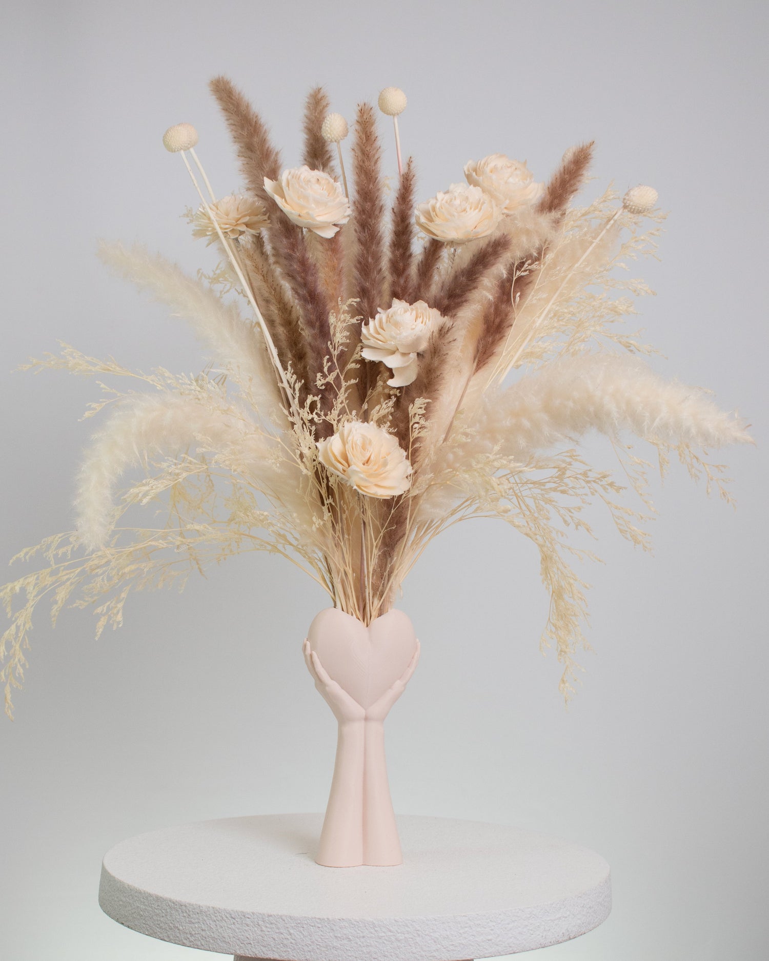Dried floral arrangement with pampas grass and preserved flowers in neutral tones, displayed in a heart-in-hand vase