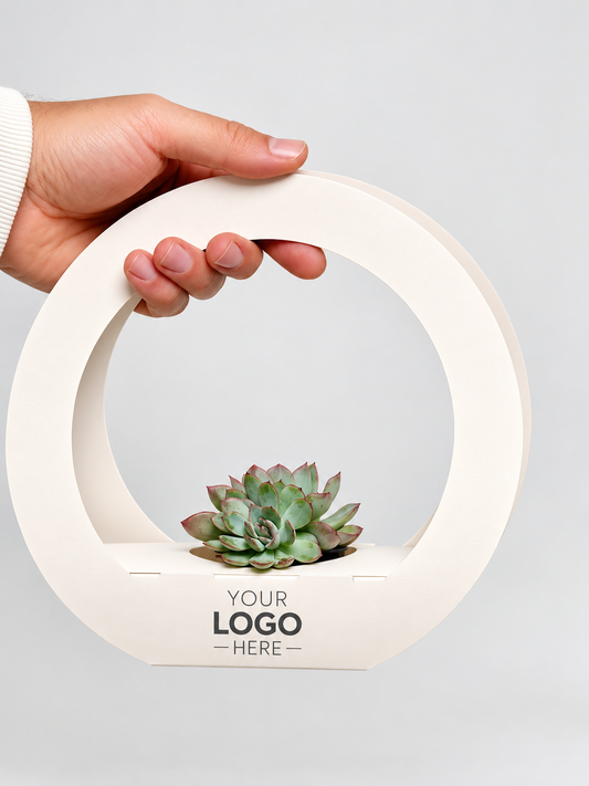 A hand holding a circular off-white wooden planter with a small succulent plant inside. The planter features a handle and a base printed with the placeholder text 'YOUR LOGO - HERE -'.