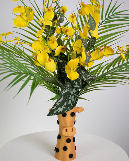 Artificial floral arrangement with bright yellow orchids and tropical leaves in a giraffe-shaped vase with black polka dots, styled on a white pedestal — a playful, tropical-inspired décor piece perfect for fun modern spaces.