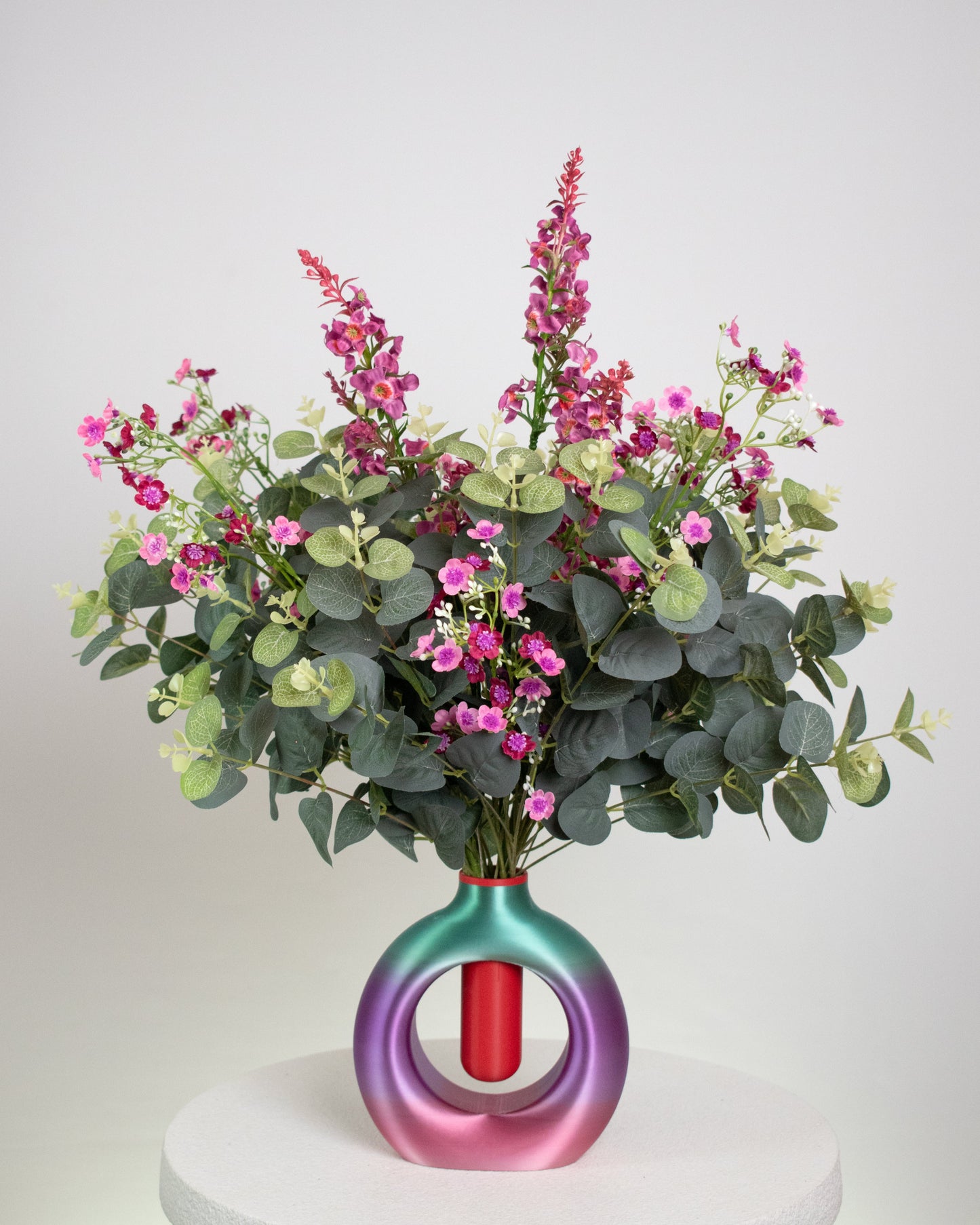 Artificial floral arrangement with pink and purple blossoms and eucalyptus greenery, displayed in a round ombre vase blending purple, green, and rose tones, on a neutral background.