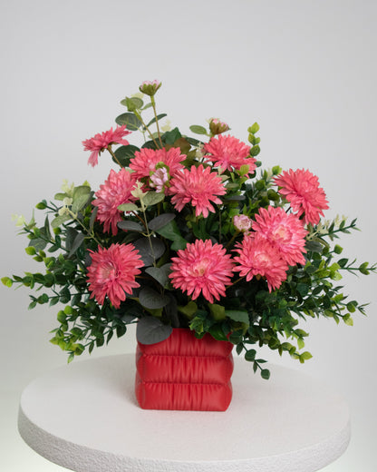 Artificial floral arrangement featuring bright pink chrysanthemums and green foliage in a soft red padded square vase, styled on a white pedestal — perfect for adding a cheerful pop of color to modern interiors.