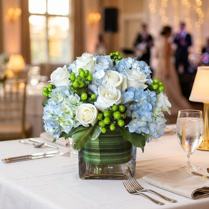 A floral arrangement featuring white and light blue hydrangeas, white roses, and green berries in a square glass vase with a green striped wrap, set on a wooden surface against a red brick wall.