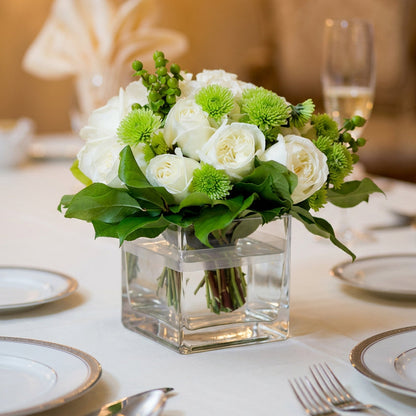 A floral centerpiece arrangement of white roses and green pom-pom chrysanthemums in a clear square glass vase, set on a white table with plates and a blurred dining background.