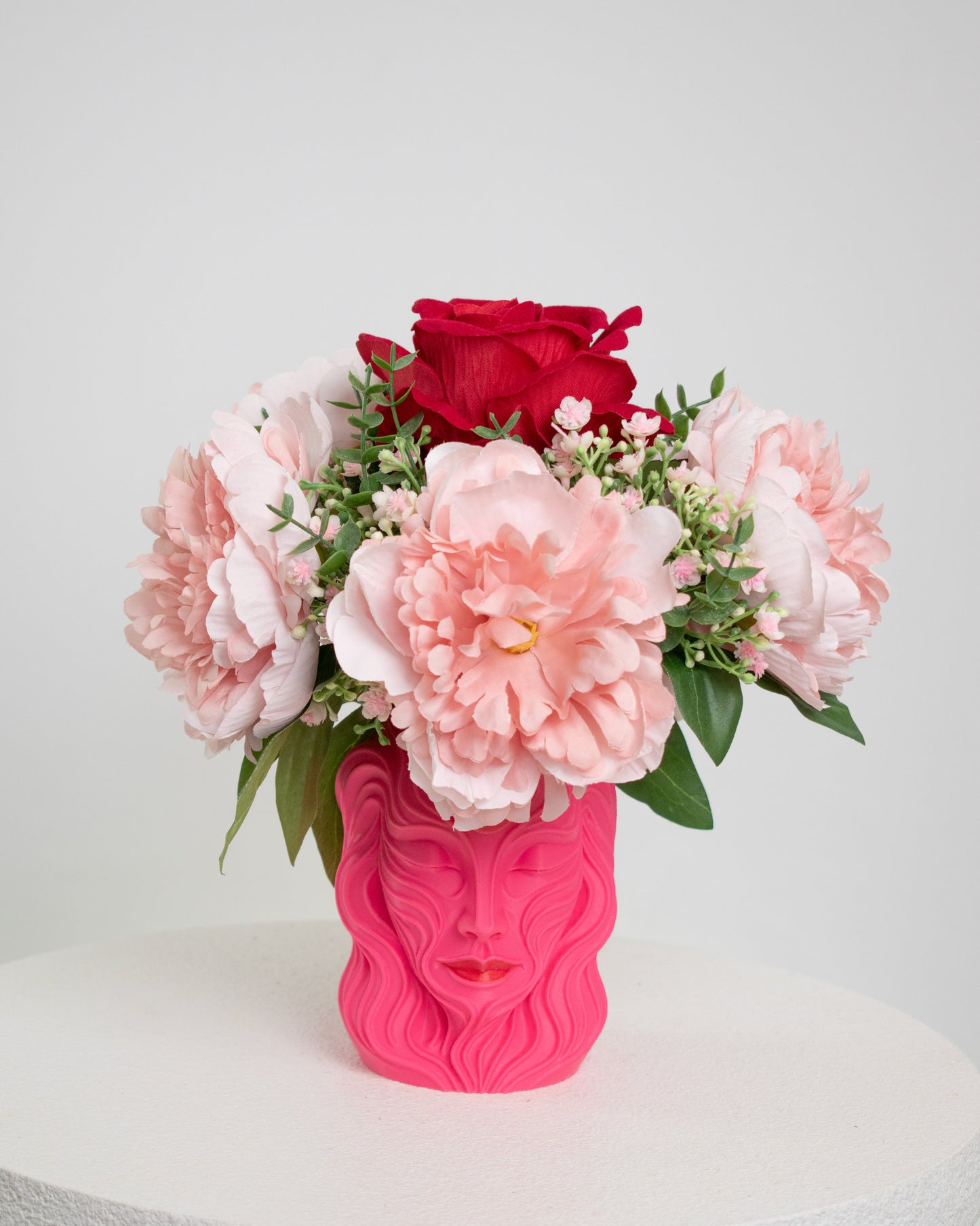 Artificial floral arrangement with blush pink peonies and a red rose in a sculpted pink vase shaped like a woman’s face, styled on a white pedestal — elegant and feminine décor for modern interiors.