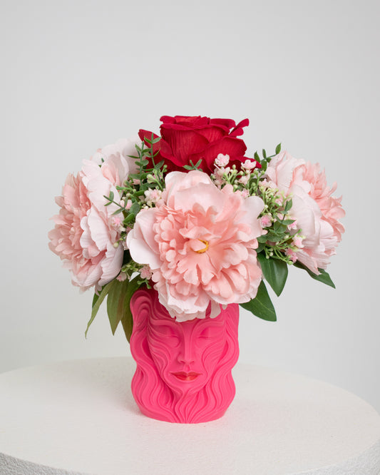 Artificial floral arrangement with blush pink peonies and a red rose in a sculpted pink vase shaped like a woman’s face, styled on a white pedestal — elegant and feminine décor for modern interiors.