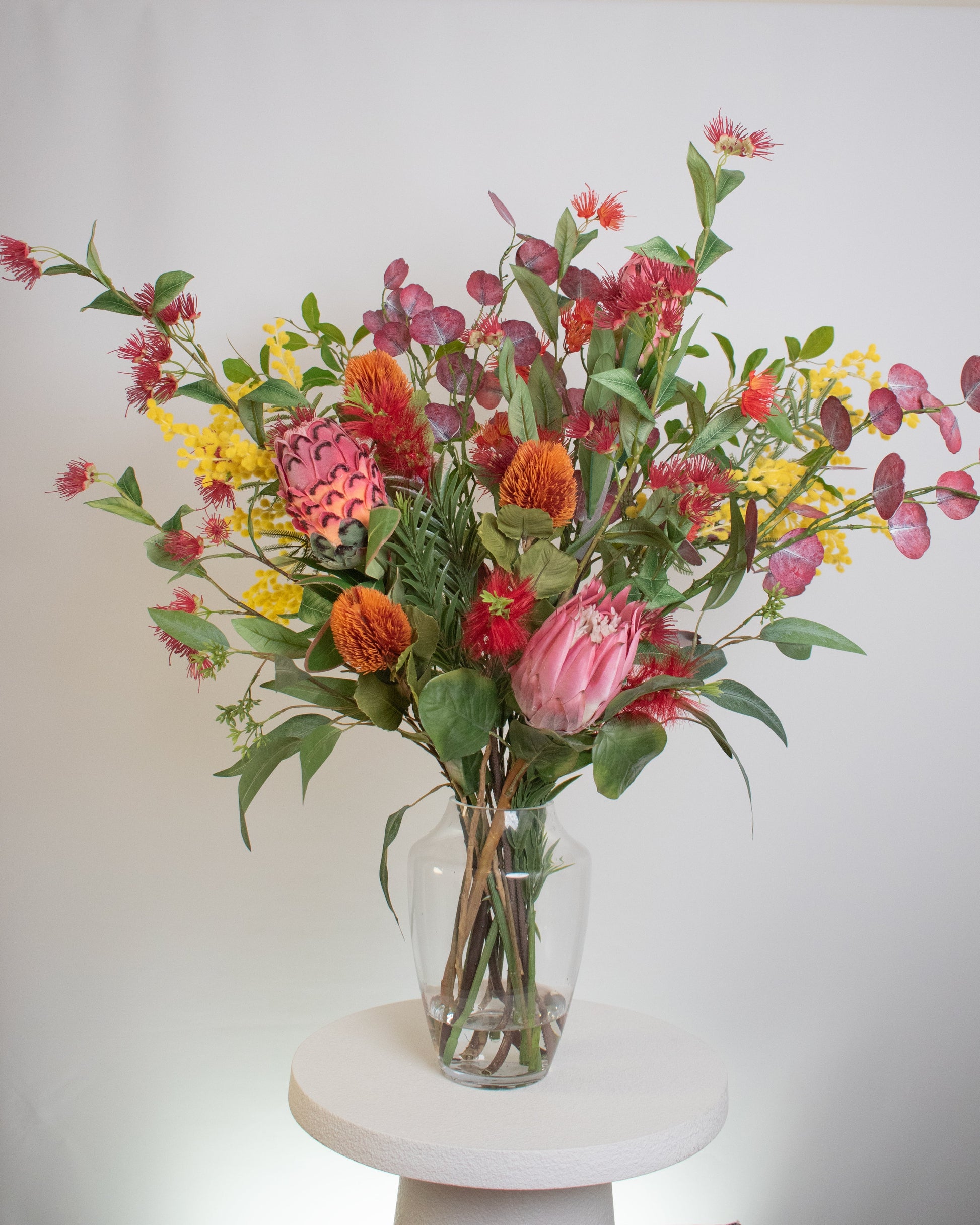 A vibrant arrangement of artificial Australian native flowers, including protea, banksia, wattle, and eucalyptus, displayed in a clear glass vase on a white pedestal.