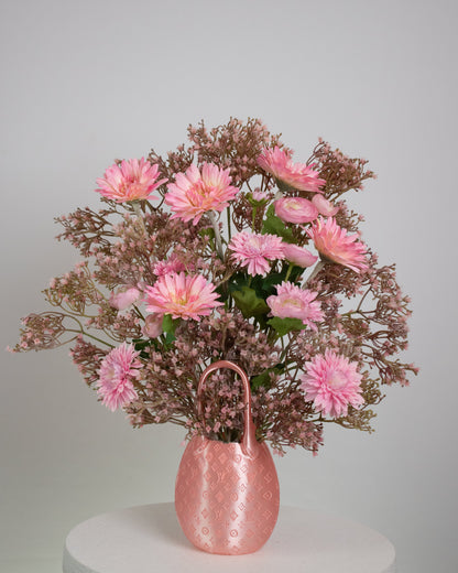 Artificial pink floral arrangement with daisies and ranunculus, accented with soft rose-toned filler, arranged in a designer-inspired pink bag vase on a neutral background.