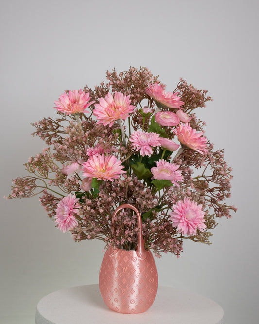 Artificial pink floral arrangement with daisies and ranunculus, accented with soft rose-toned filler, arranged in a designer-inspired pink bag vase on a neutral background.
