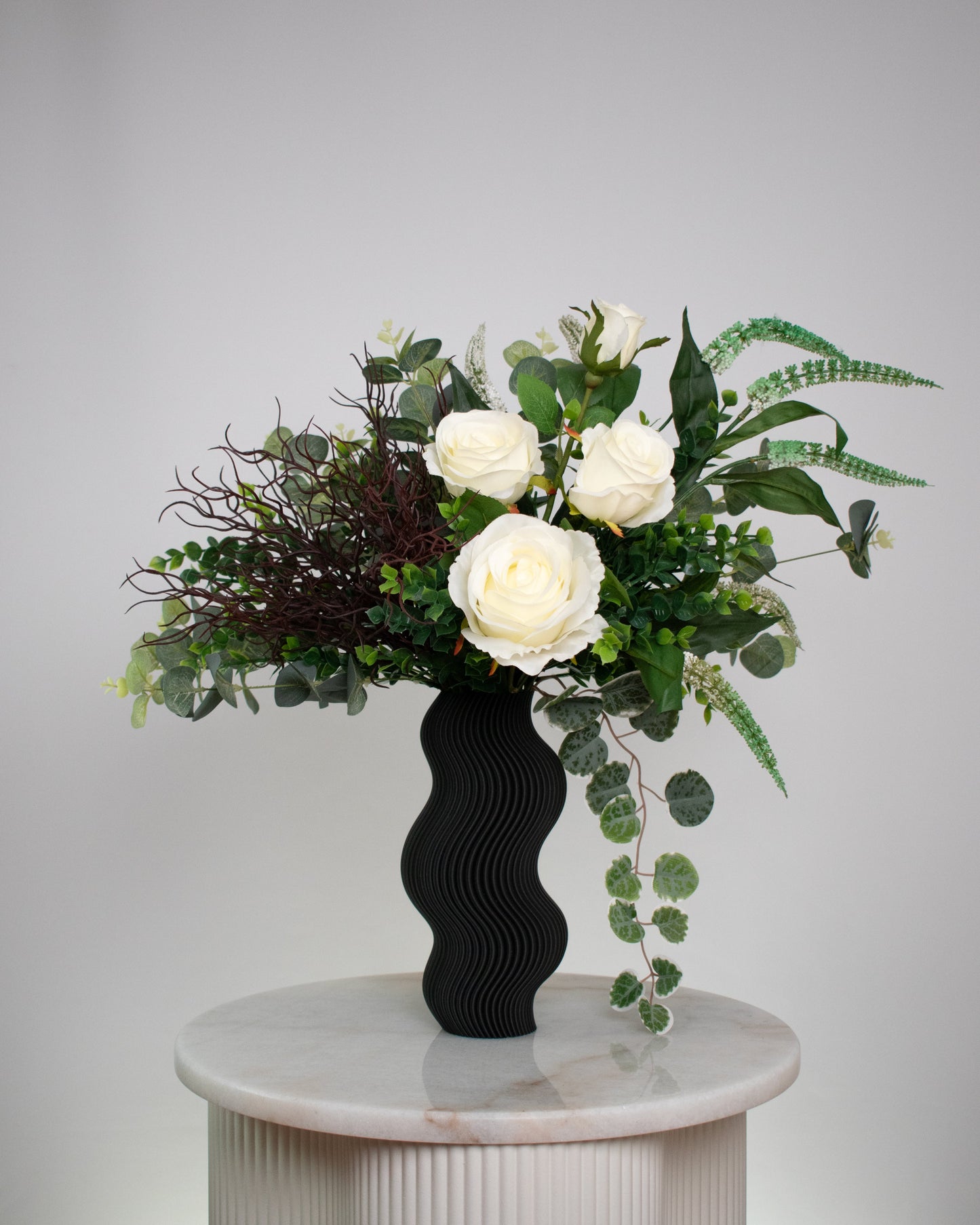 Artificial white rose arrangement with lush greenery in a sculptural matte black wavy vase, styled on a marble table — a modern and elegant decorative floral display ideal for minimalist interiors.