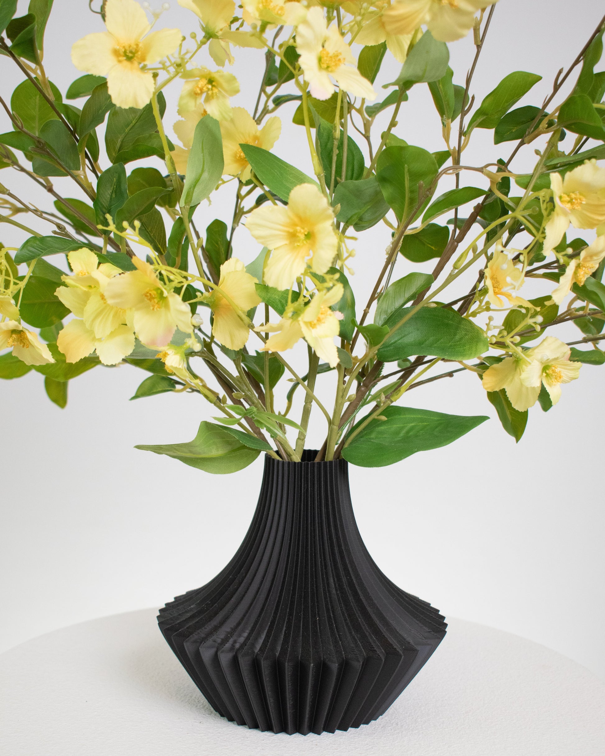 Artificial yellow floral arrangement with lush greenery in a modern black pleated vase