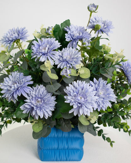 Artificial floral arrangement with soft lavender-blue flowers and lush green leaves in a puffed satin-blue vase, styled on a white pedestal against a neutral background.
