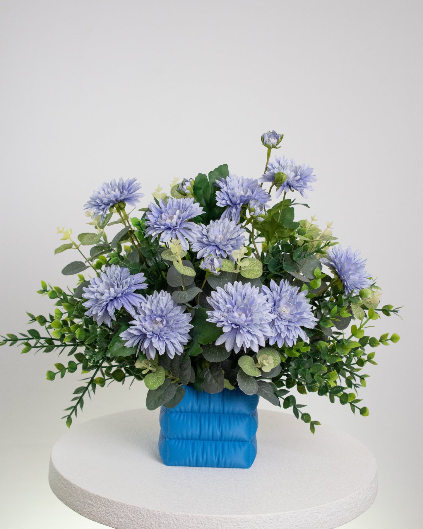 Artificial floral arrangement with soft lavender-blue flowers and lush green leaves in a puffed satin-blue vase, styled on a white pedestal against a neutral background.
