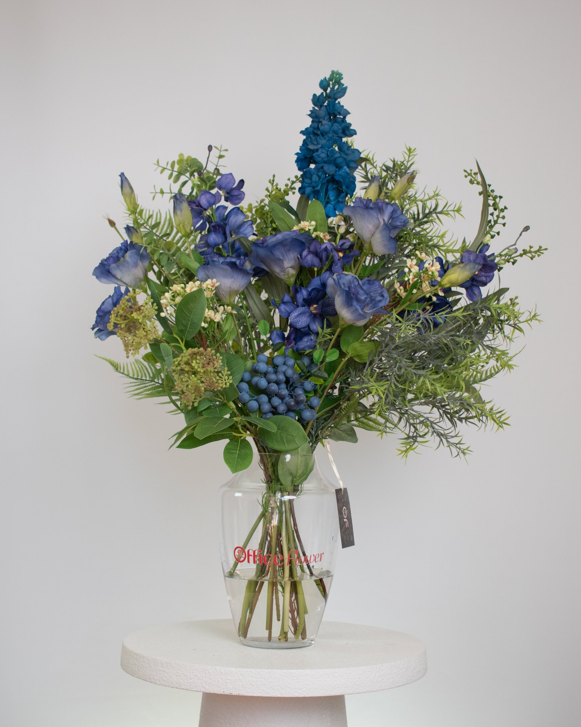 Artificial blue floral arrangement with greenery and berry accents, displayed in a clear glass vase with acrylic water