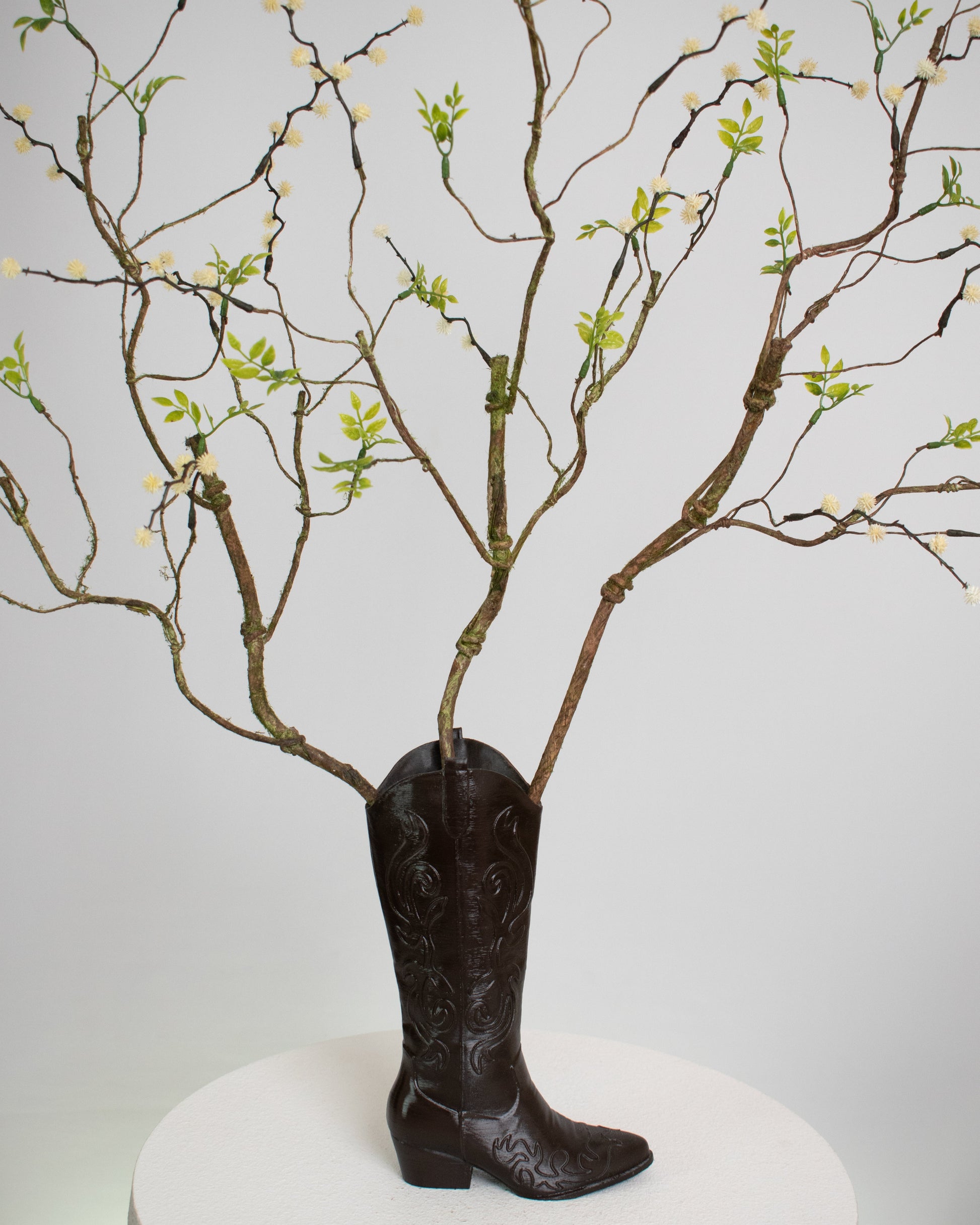 Rustic artificial floral arrangement with natural branches and light green leaves arranged in a dark brown cowboy boot-shaped vase, styled as a modern rustic décor accent.