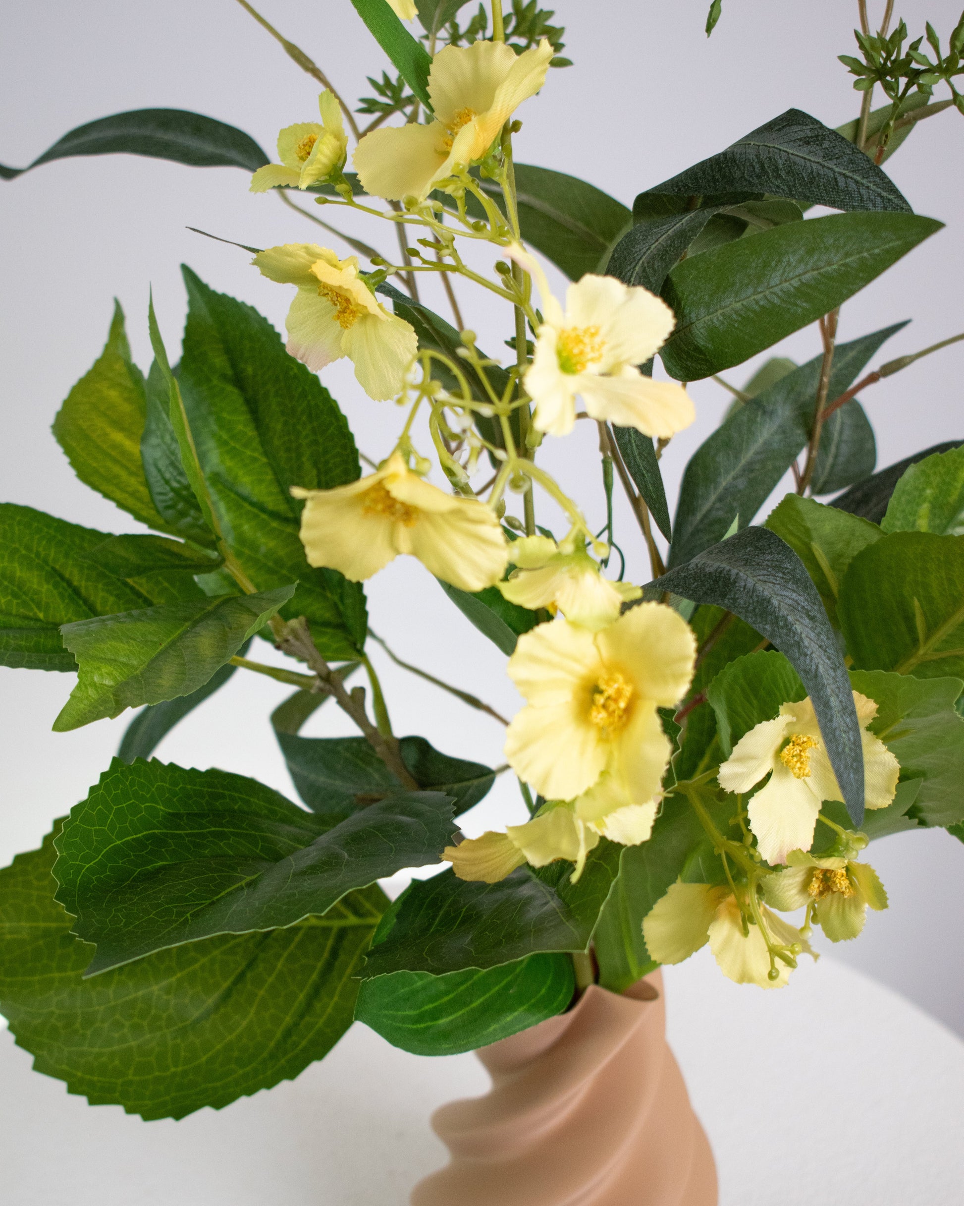 Artificial floral arrangement featuring soft yellow flowers and green leaves in a twisted matte caramel vase, displayed on a white round pedestal against a neutral background.