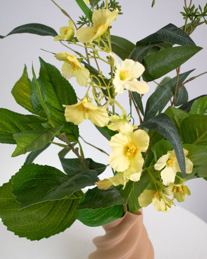 Artificial floral arrangement featuring soft yellow flowers and green leaves in a twisted matte caramel vase, displayed on a white round pedestal against a neutral background.