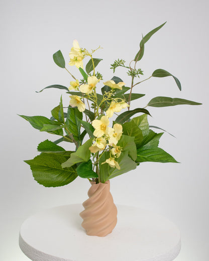 Artificial floral arrangement featuring soft yellow flowers and green leaves in a twisted matte caramel vase, displayed on a white round pedestal against a neutral background.