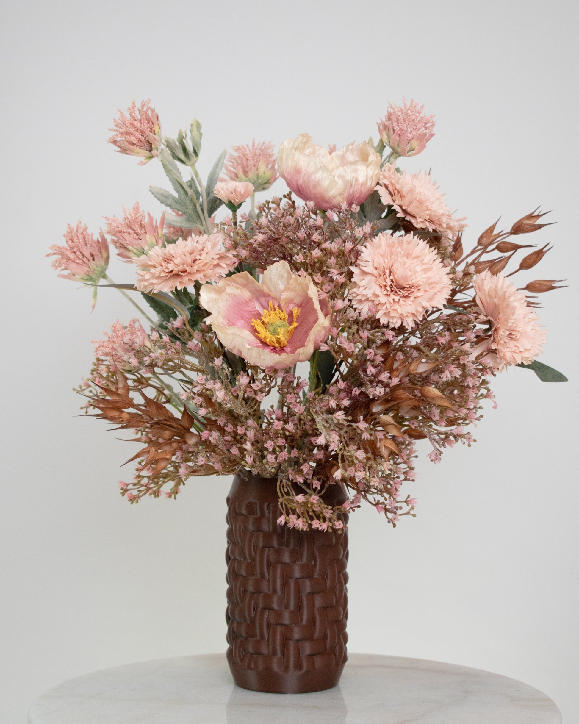 Artificial floral arrangement with blush pink flowers and rustic brown foliage in a woven brown vase, styled on a marble table — a warm, elegant decorative piece perfect for neutral or earthy interiors.