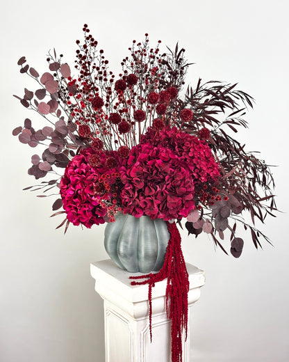 Decorative floral arrangement with red flowers and greenery in a gray vase on a white pedestal against a light gray background.