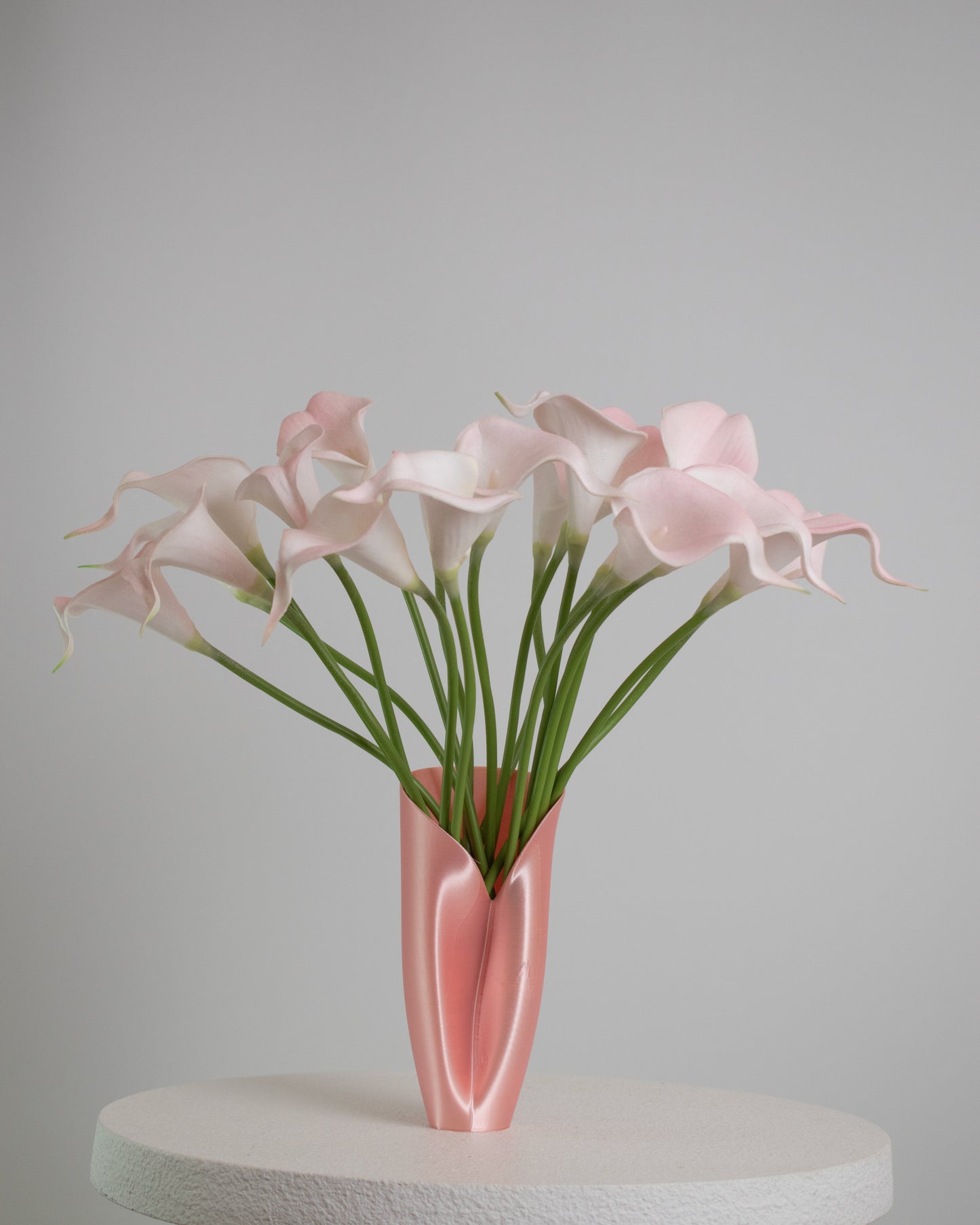Artificial floral arrangement of soft pink calla lilies with lifelike green stems, elegantly displayed in a sleek blush pink vase on a neutral background