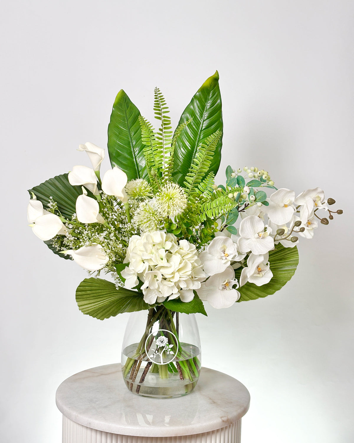A white and green floral arrangement in a clear glass vase, displayed on a round white marble pedestal against a light gray background.