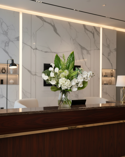 Floral arrangement for a modern reception desk in a luxury lobby with a dark wood grain front, gold trim, and a marble-patterned back wall. A vibrant floral arrangement of white flowers and green leaves sits in the center of the desk