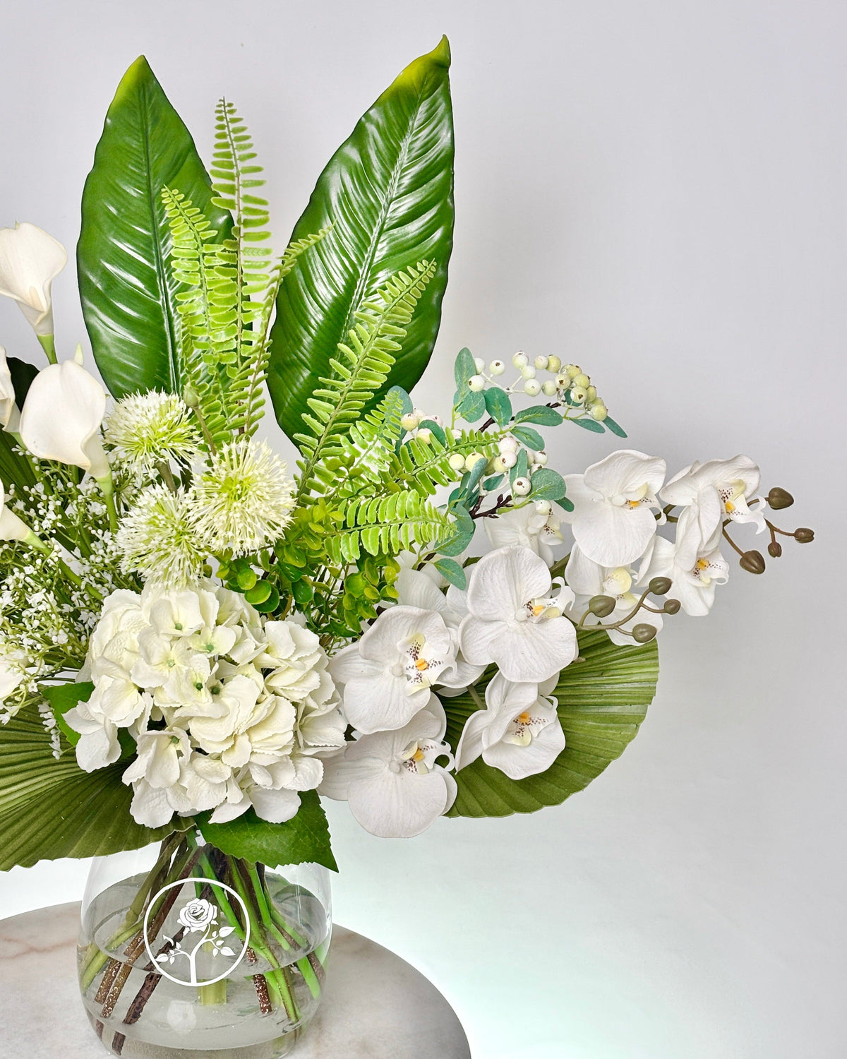 A lush faux floral arrangement in a clear glass vase, featuring white orchids, white hydrangeas, green ferns, and large green tropical leaves, displayed on a light marble surface against a plain light gray background.