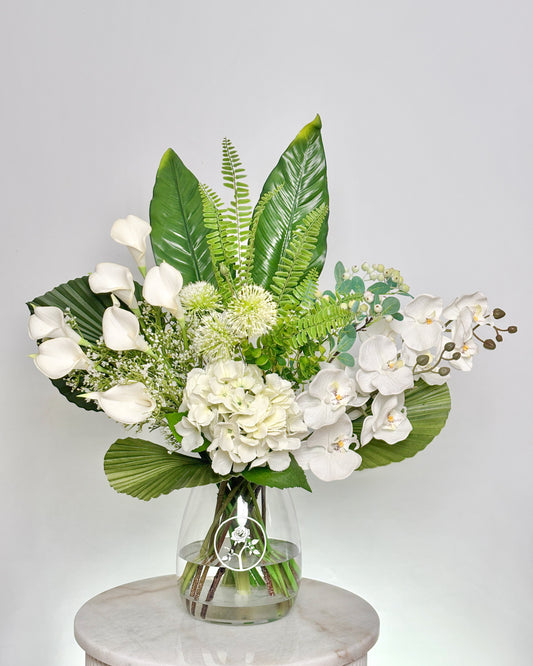 A lush floral arrangement featuring white calla lilies, white orchids, green tropical leaves, and ferns in a clear glass vase, displayed on a white marble pedestal against a light gray background.
