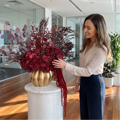 A woman in a beige blouse and navy trousers stands beside a large dried floral arrangement in a gold vase on a white pedestal inside a modern office space.
