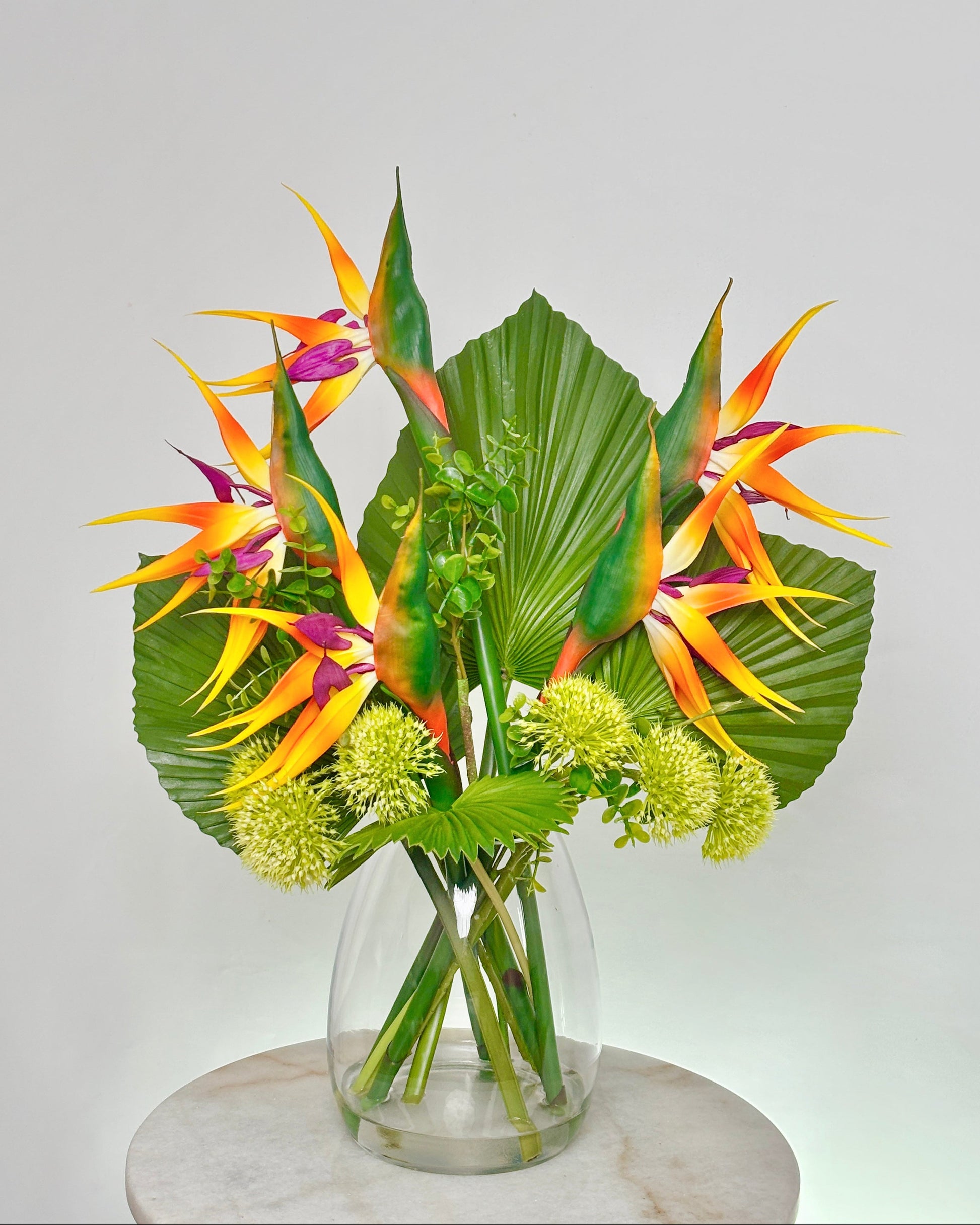 A tropical floral arrangement featuring vibrant bird of paradise flowers, large green palm leaves, and small pale yellow globe flowers in a clear glass vase, displayed on a round white marble pedestal against a light gray background