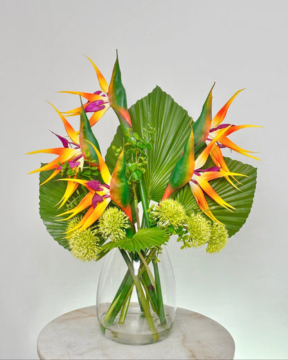 A tropical floral arrangement featuring vibrant bird of paradise flowers, large green palm leaves, and small pale yellow globe flowers in a clear glass vase, displayed on a round white marble pedestal against a light gray background