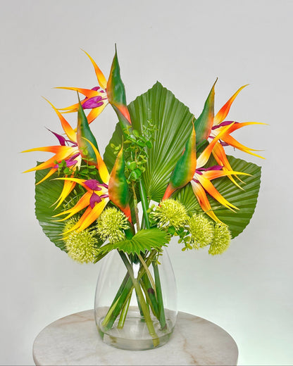 A tropical artificial floral arrangement featuring vibrant bird of paradise flowers, large green palm leaves, and small pale yellow globe flowers in a clear glass vase, displayed on a round white marble pedestal against a light gray background.