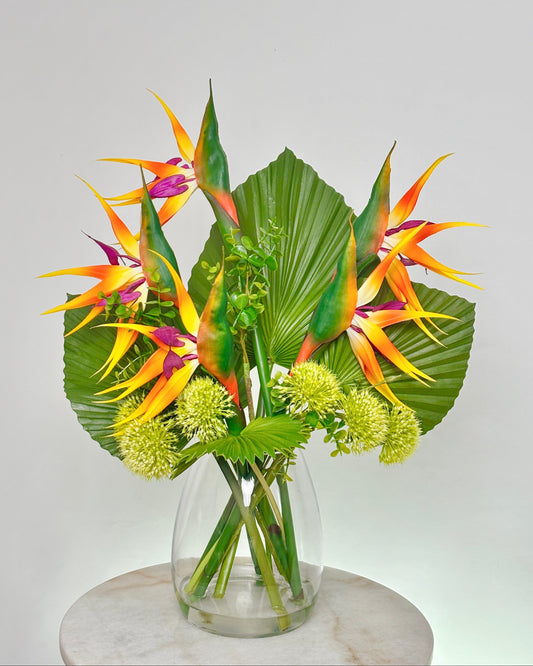 A tropical artificial floral arrangement featuring vibrant bird of paradise flowers, large green palm leaves, and small pale yellow globe flowers in a clear glass vase, displayed on a round white marble pedestal against a light gray background.