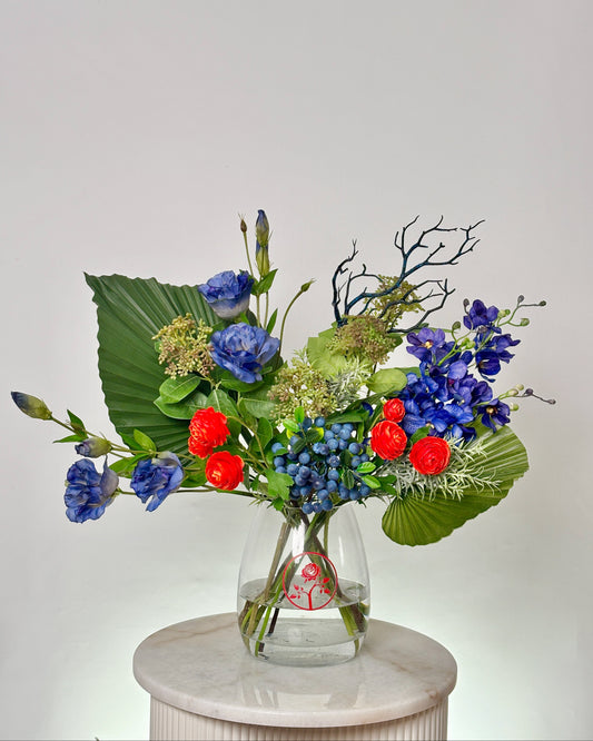 A vibrant floral arrangement featuring blue flowers, red berries, green foliage, and blue-green branches, presented in a clear glass vase on a white marble pedestal against a light gray background