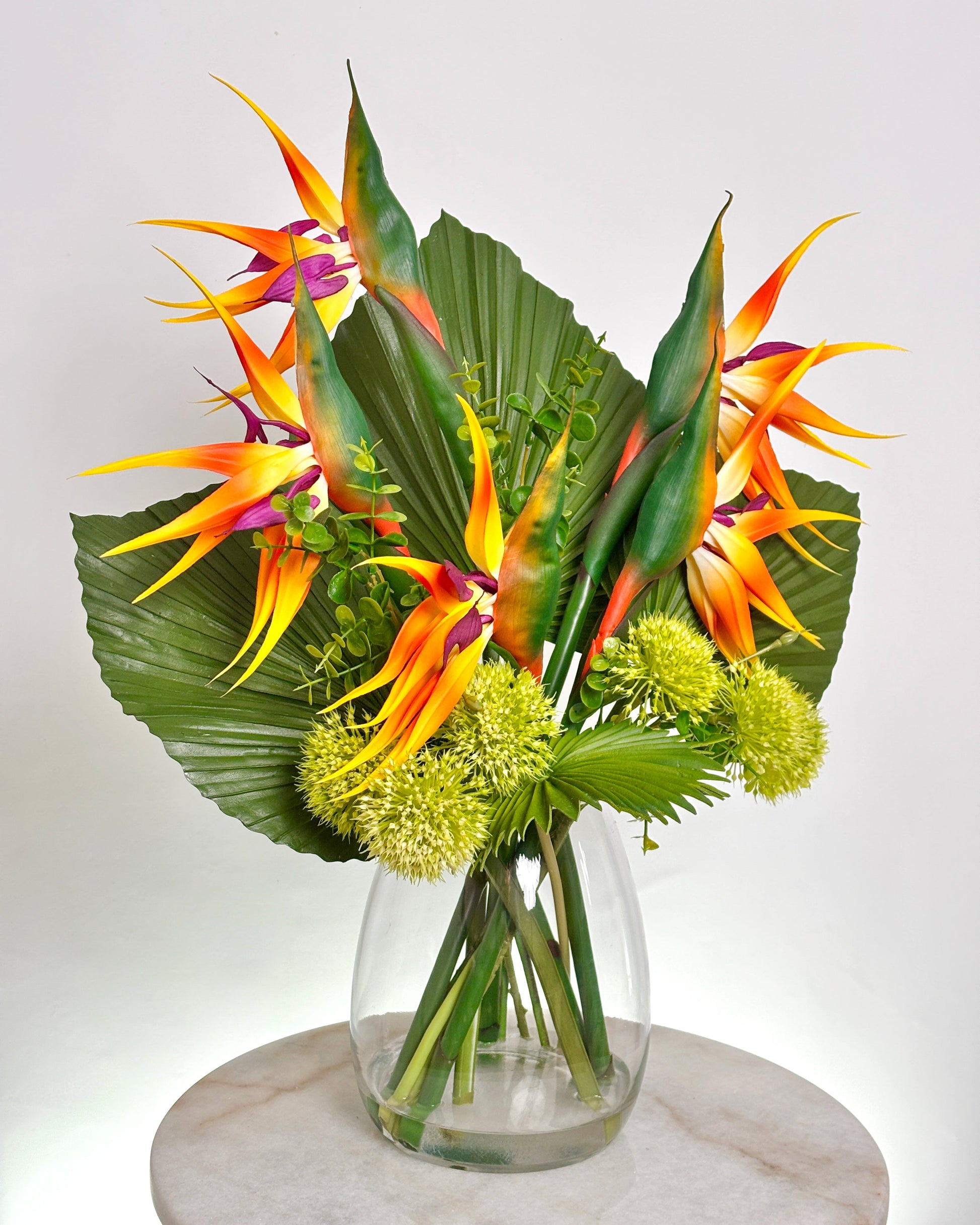 A tropical office floral arrangement featuring vibrant bird of paradise flowers, large green palm leavs, and round yellow-green globe flowers in a clear glass vase, displayed on a round marble surface against a light gray background.