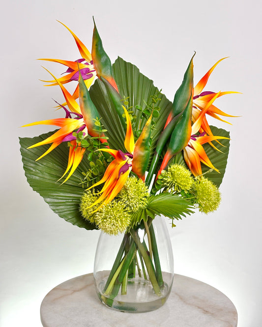A tropical office floral arrangement featuring vibrant bird of paradise flowers, large green palm leavs, and round yellow-green globe flowers in a clear glass vase, displayed on a round marble surface against a light gray background.