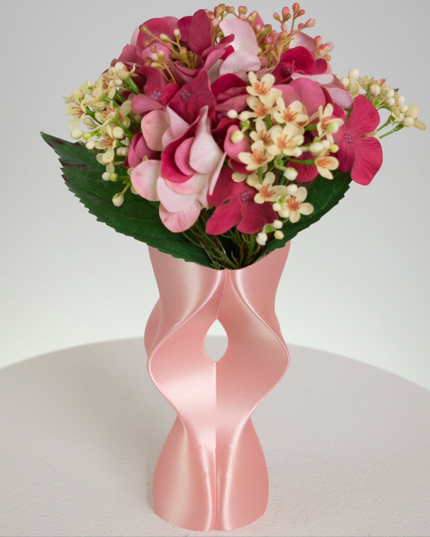 Artificial pink floral arrangement featuring rosy hydrangeas and cream blossoms, beautifully displayed in a sculptural modern pink vase with curved design.