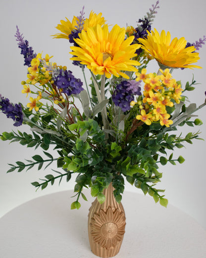 Artificial flower arrangement featuring bright yellow gerberas, purple lavender sprigs, and lush greenery in a golden vase embossed with a sunflower design, ideal for warm or rustic interiors.