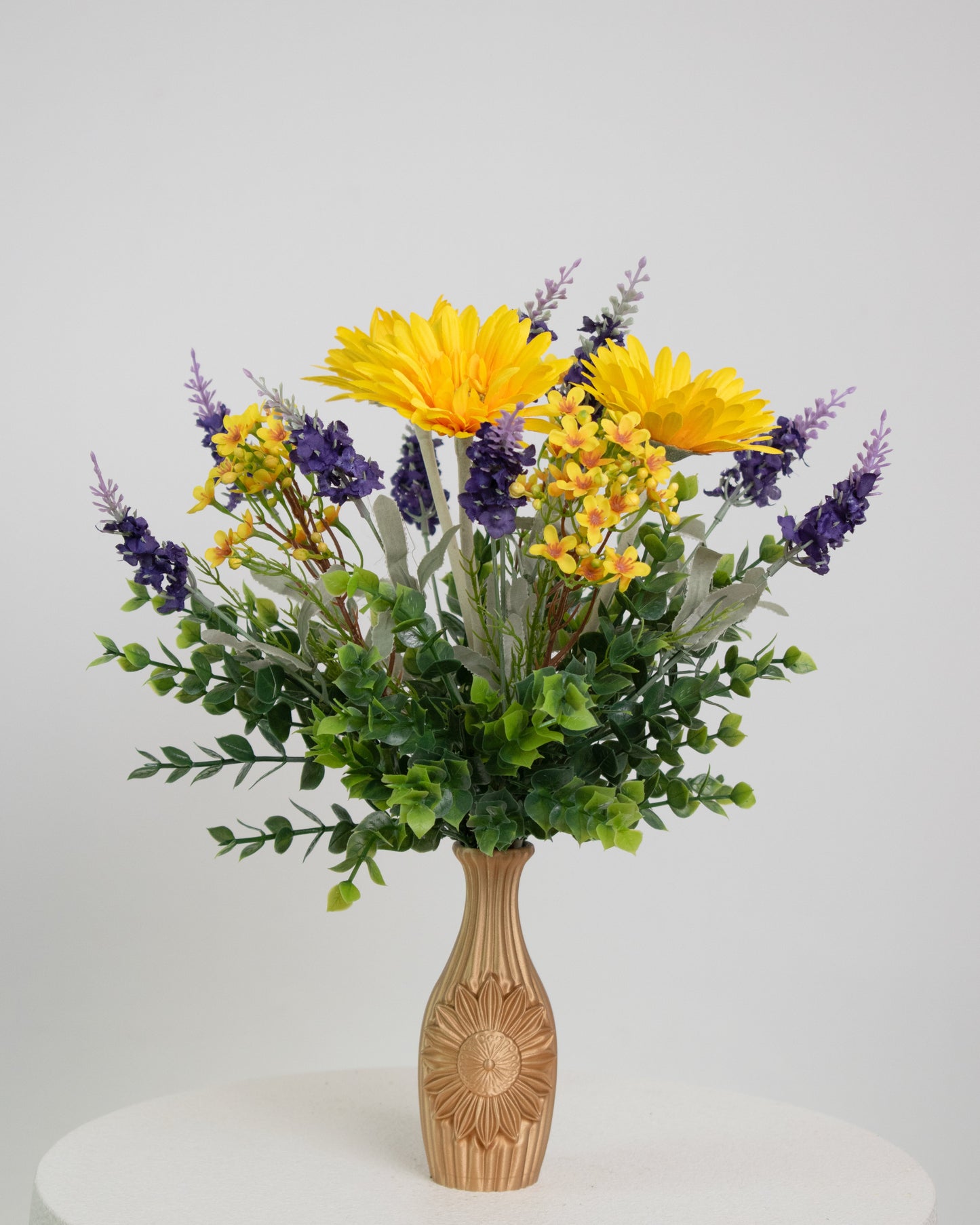 Artificial flower arrangement featuring bright yellow gerberas, purple lavender sprigs, and lush greenery in a golden vase embossed with a sunflower design, ideal for warm or rustic interiors.