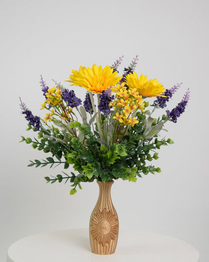 Artificial flower arrangement featuring bright yellow gerberas, purple lavender sprigs, and lush greenery in a golden vase embossed with a sunflower design, ideal for warm or rustic interiors.