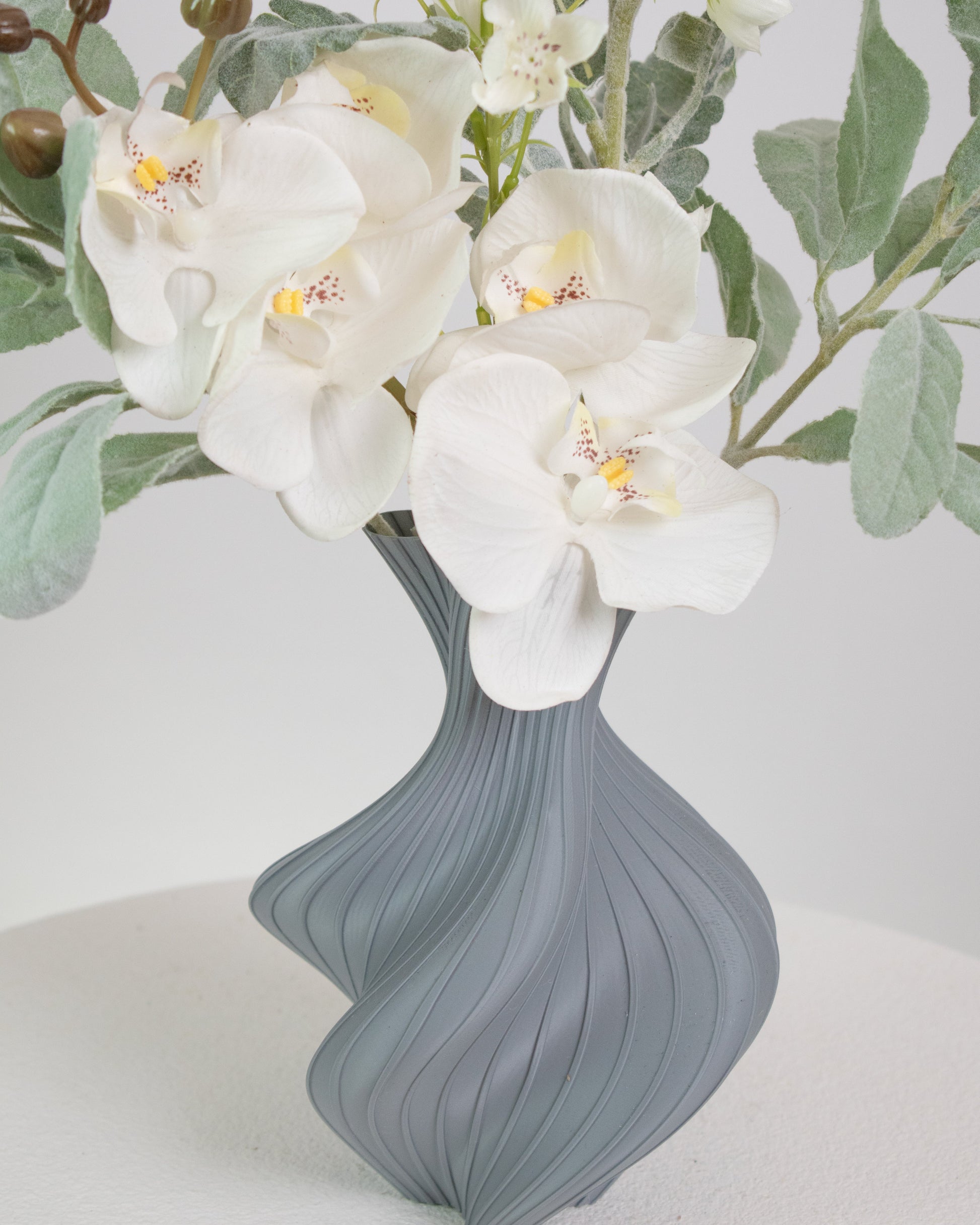 Artificial floral arrangement featuring white orchids and silver-green foliage in a sculptural grey vase with curved lines, displayed on a round white pedestal.