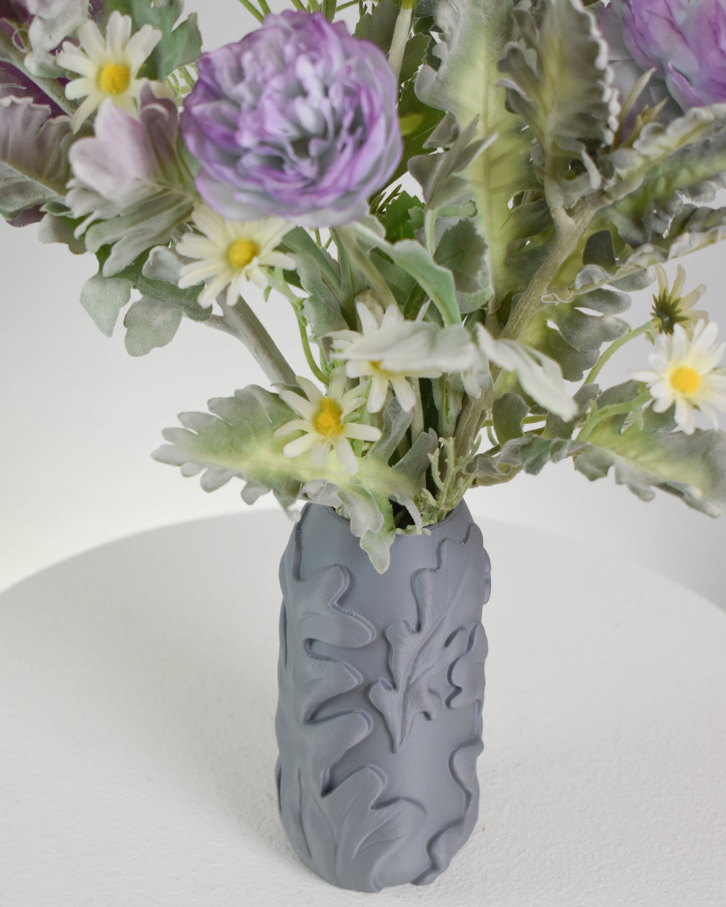 Artificial floral arrangement featuring lavender blooms, white daisies, and green foliage, displayed in a matte grey textured vase on a neutral background.