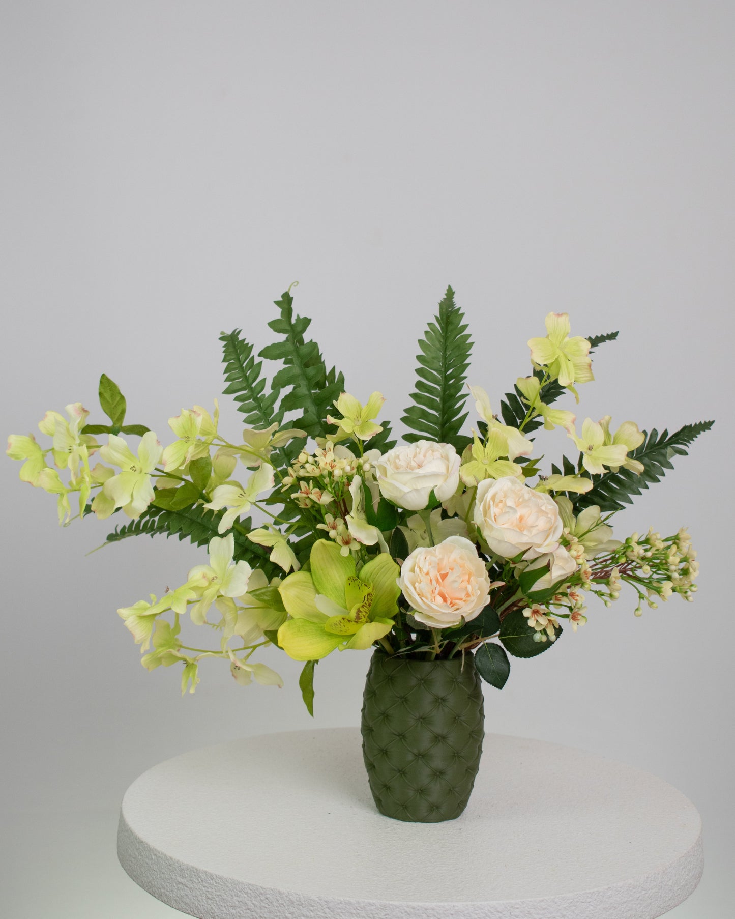 Artificial floral arrangement featuring cream roses, green orchids, and lush ferns in a textured olive-green vase, styled on a white pedestal — perfect for adding a refreshing, natural touch to modern or earthy-toned interiors.