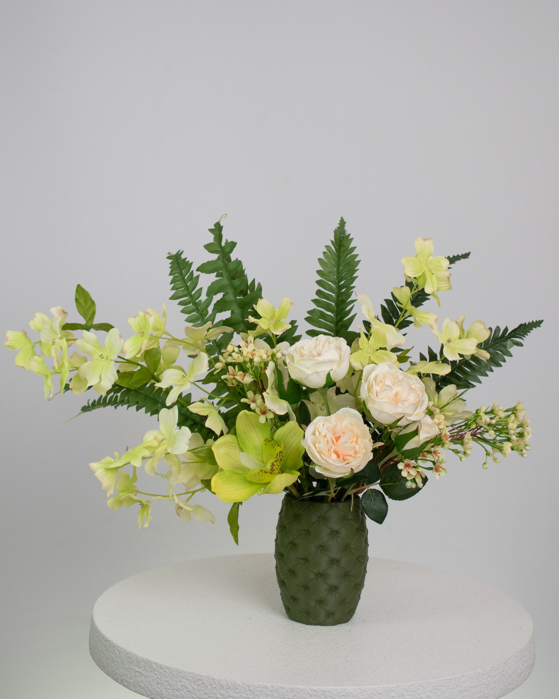 Artificial floral arrangement featuring cream roses, green orchids, and lush ferns in a textured olive-green vase, styled on a white pedestal — perfect for adding a refreshing, natural touch to modern or earthy-toned interiors.