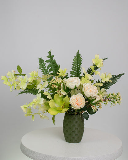 Artificial floral arrangement featuring cream roses, green orchids, and lush ferns in a textured olive-green vase, styled on a white pedestal — perfect for adding a refreshing, natural touch to modern or earthy-toned interiors.