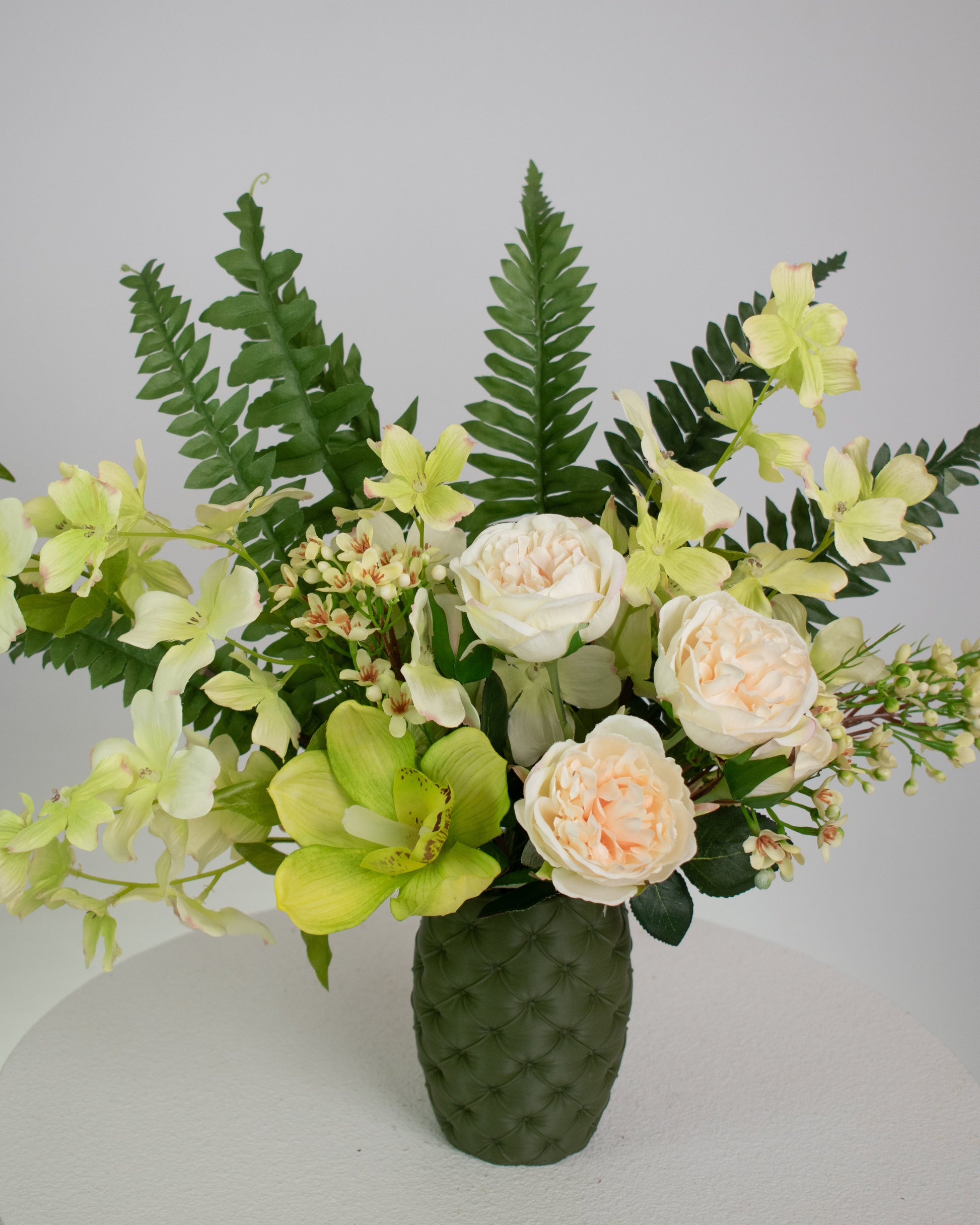 Artificial floral arrangement featuring cream roses, green orchids, and lush ferns in a textured olive-green vase, styled on a white pedestal — perfect for adding a refreshing, natural touch to modern or earthy-toned interiors.