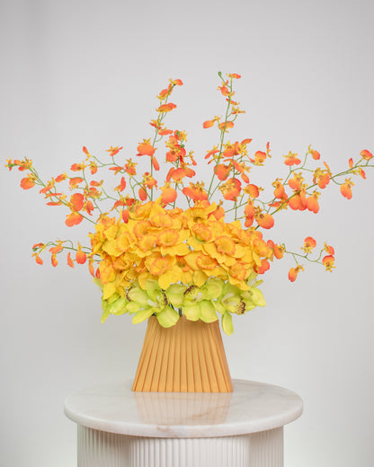 Vibrant artificial floral arrangement featuring orange orchids, yellow blossoms, and green cymbidiums in a pleated terracotta vase, styled on a white pedestal against a soft neutral background.