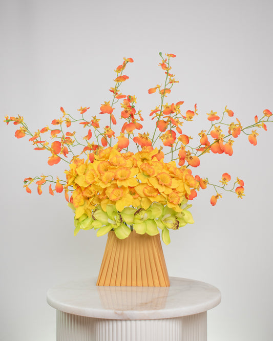 Vibrant artificial floral arrangement featuring orange orchids, yellow blossoms, and green cymbidiums in a pleated terracotta vase, styled on a white pedestal against a soft neutral background.