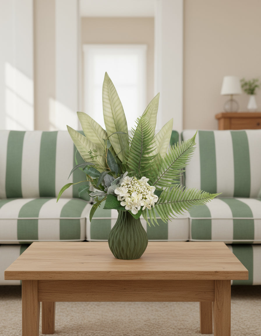 Artificial green arrangement featuring tropical leaves, ferns, eucalyptus, and ivory blooms, arranged in a textured olive-green vase on a neutral background.