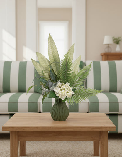 Artificial green arrangement featuring tropical leaves, ferns, eucalyptus, and ivory blooms, arranged in a textured olive-green vase on a neutral background.